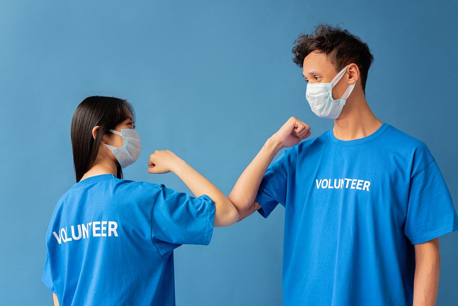 Ação Comunitária. — Two volunteers wearing blue shirts and face masks elbow bump for safety.